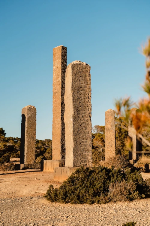 Tall stone pillars stand against a clear blue sky, surrounded by sparse vegetation. The scene evokes a sense of ancient history and isolation.