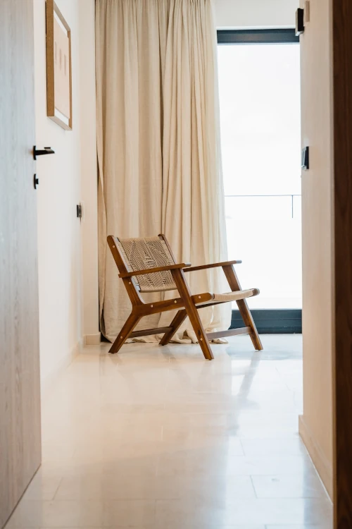 A wooden chair with woven backrest sits in a sunlit room, facing large curtains and a bright window. The setting is minimal, creating a serene atmosphere.