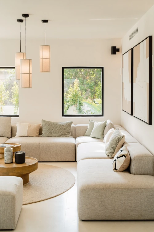 A modern living room with a beige sectional sofa and assorted pillows. Pendant lights hang above a round wooden coffee table. Large windows offer a garden view.