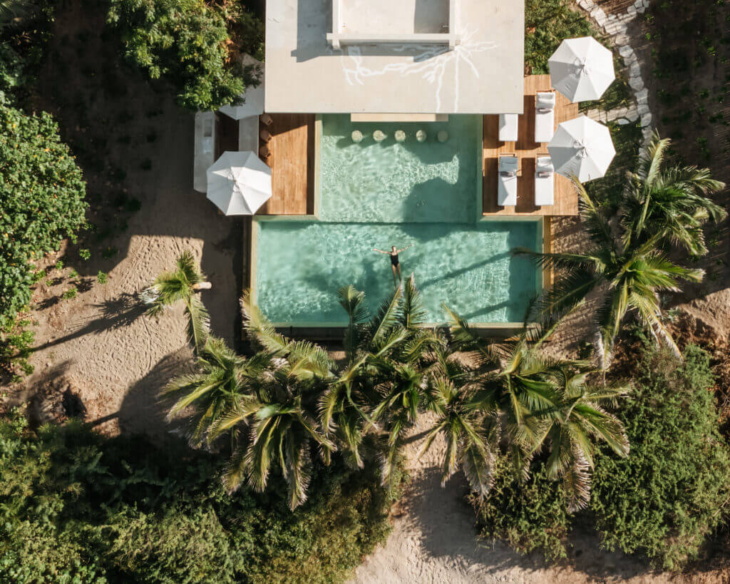 Aerial view of a villa pool with a guest floating in the water, surrounded by palms and shaded loungers.