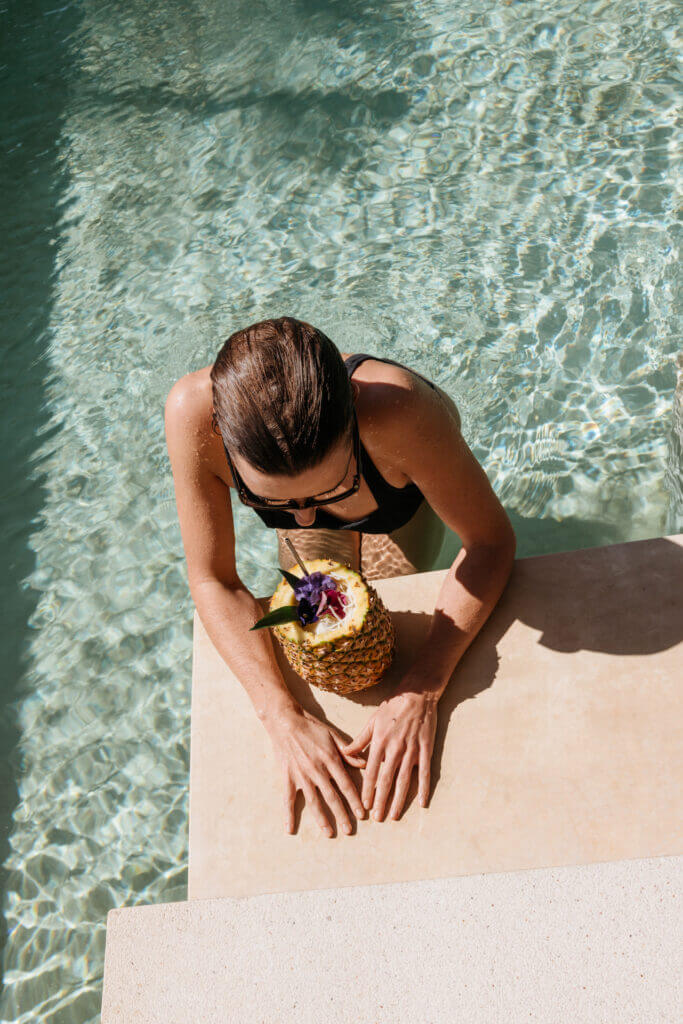 Guest in the pool beside a pineapple-shaped drink garnished with flowers.
