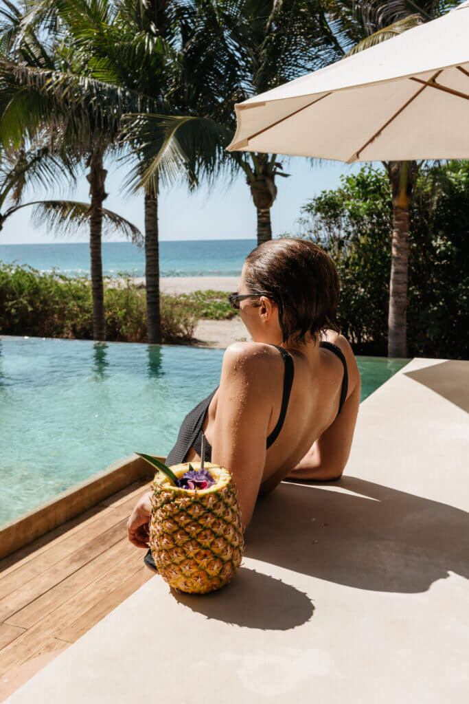 Guest relaxing beside the infinity pool with a pineapple drink and ocean view.