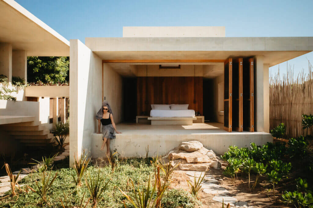 Open-air bedroom with a low bed and a guest seated on the villa’s stone terrace.