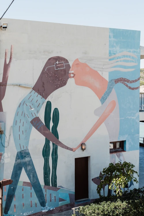 A vibrant mural on a building wall depicts two abstract figures leaning in for a kiss, set against a light background. A cactus and a few plants add detail.