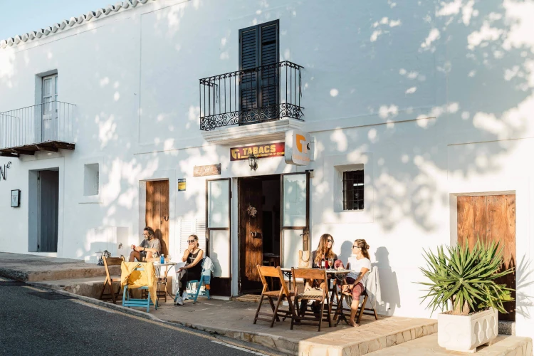 Sunlit street scene with people sitting at tables outside a white building marked 