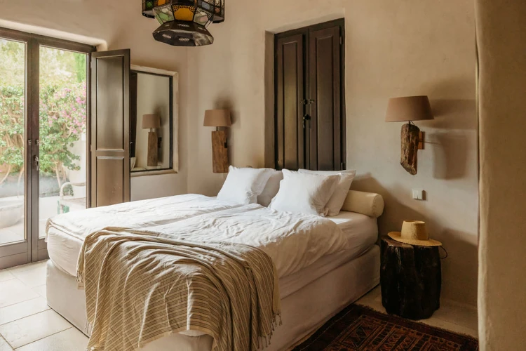 A cozy bedroom with a neatly made double bed, draped with a striped blanket. Warm lighting, rustic decor, and an open door reveal a lush garden view.