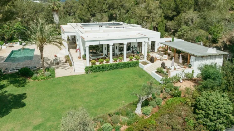 A modern white villa surrounded by lush greenery and situated beside a spacious green lawn. A rectangular pool is visible to the left, creating a serene outdoor retreat.