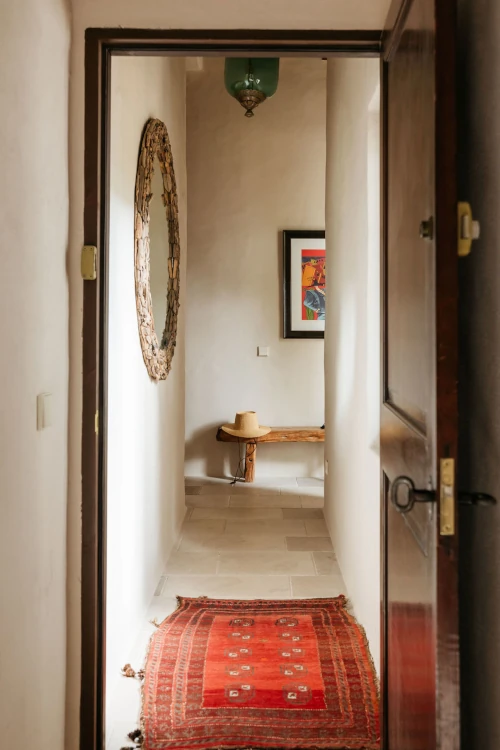 A rustic hallway with a red patterned carpet leads to a wooden bench and a colorful framed artwork. The ambiance is warm and inviting.