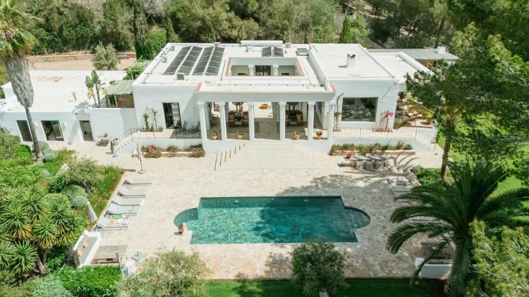 Aerial view of a modern white villa with solar panels, surrounded by lush greenery. Features a spacious patio and a rectangular swimming pool.