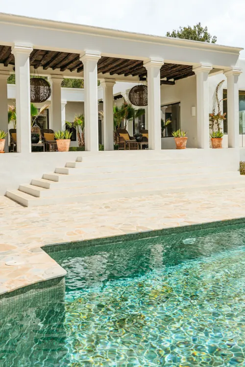 Elegant poolside villa with white columns and terra-cotta pots, lush plants, wicker furniture, and a tranquil turquoise pool. Bright, serene atmosphere.