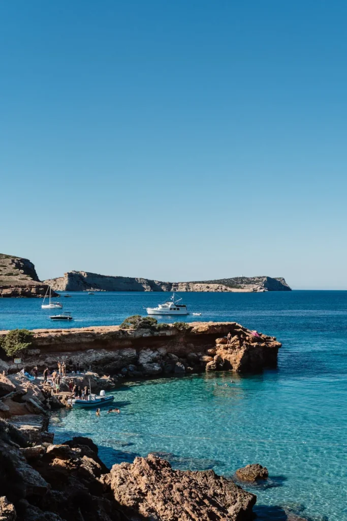 Coastal scene with clear blue sea, rocky cliffs, and several anchored boats. People gather on the shore, enjoying a sunny day. The mood is tranquil.