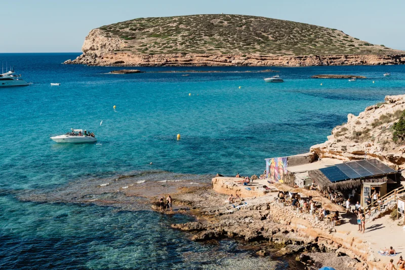 Rocky beach with a small bar and people sunbathing. Turquoise sea features boats with a large island in the background under clear blue sky.