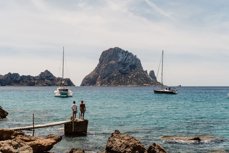 Two people stand on a dock by the sea, facing calm turquoise water with two anchored sailboats. A rocky island looms in the distance under a blue sky.