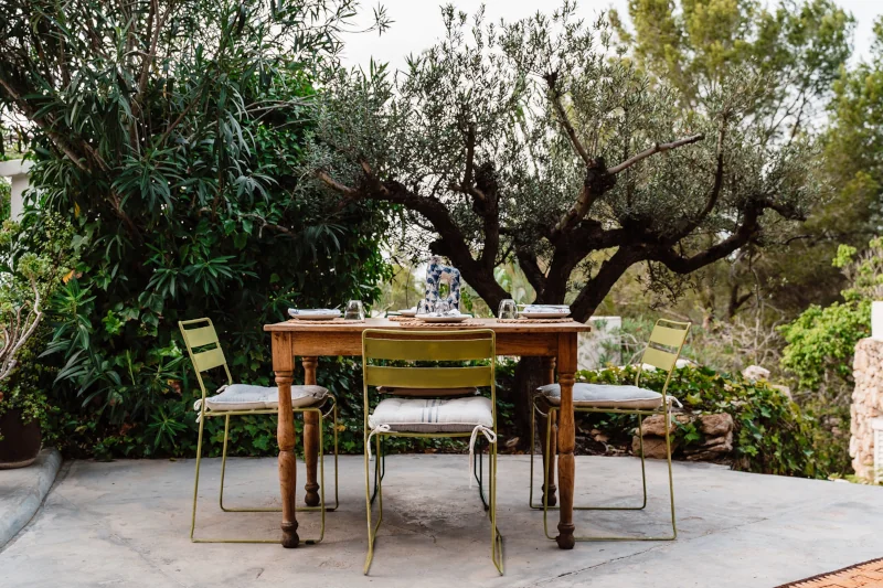 Outdoor setting with a rustic wooden table, surrounded by four green chairs. The table is set with dishes and placed near lush greenery and trees, conveying a peaceful, natural ambiance.