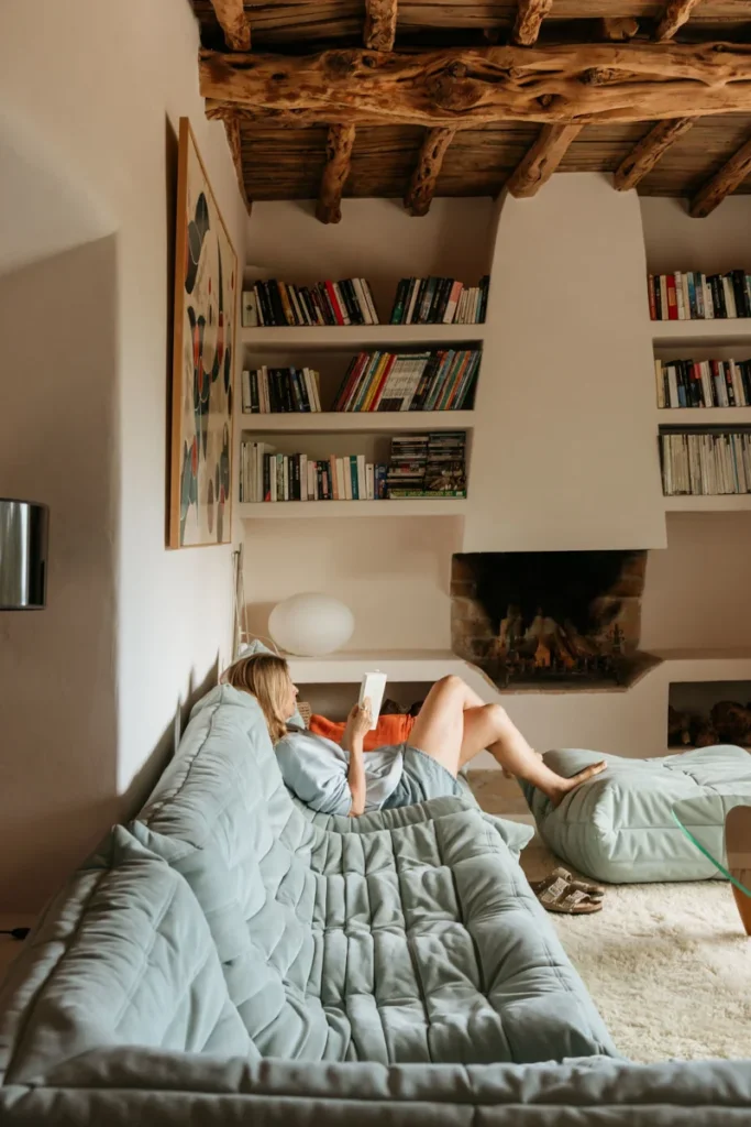 A person relaxes on a plush, light blue sofa, reading a book in a cozy living room. Wooden beams, bookshelves, and a lit fireplace create a warm ambiance.