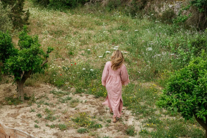 A woman in a long pink dress walks through a lush, flower-filled garden. The scene is tranquil and serene, with green trees framing the path.