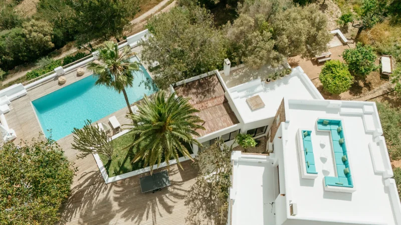 Aerial view of a modern villa with a turquoise pool, surrounded by lush greenery. White architecture and palm trees create a serene, luxurious ambiance.