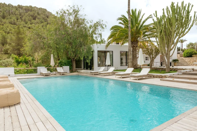 Luxurious outdoor pool with clear blue water surrounded by white lounge chairs and umbrellas. Lush greenery, palm trees, and a modern white house create a serene, tropical ambiance.