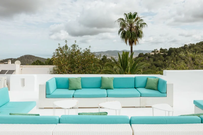 Contemporary patio with turquoise sofas, white tables, and lush greenery. A palm tree stands under a cloudy sky, creating a serene, tropical ambiance.