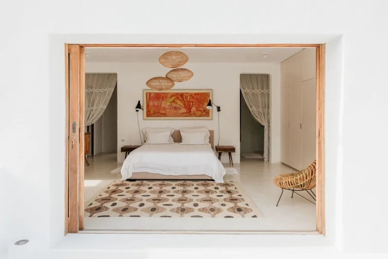 Minimalist bedroom with a large window view. Features a white bed, wooden decor, earthy tones, patterned rug, and woven chairs, conveying tranquility.