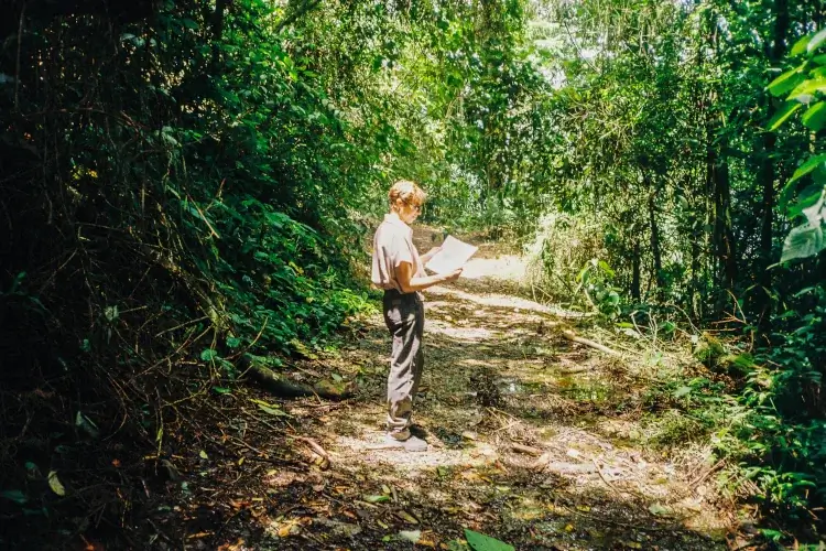 A person in hiking gear standing on a sun-dappled dirt trail and reviewing a map, surrounded by dense, green tropical foliage and towering trees at Hacienda Oro Molido.