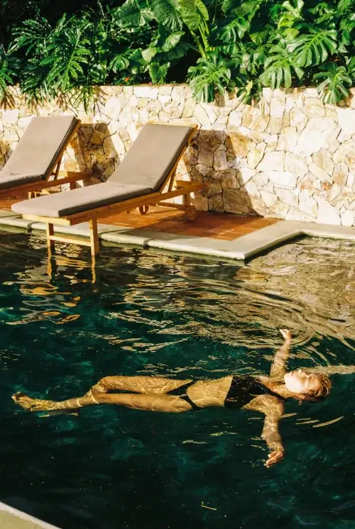 A woman in a black bikini floating peacefully in a clear swimming pool at Hacienda Oro Molido, with wooden lounge chairs, a stone wall, and tropical Monstera plants in the background.