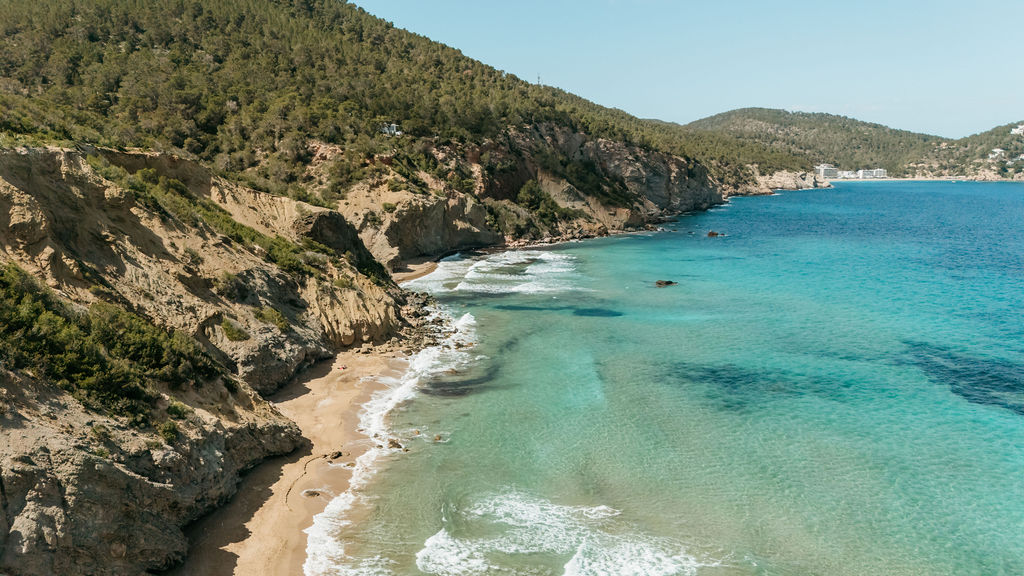 Turquoise waves break along a secluded cove beneath Ibiza’s wooded cliffs.