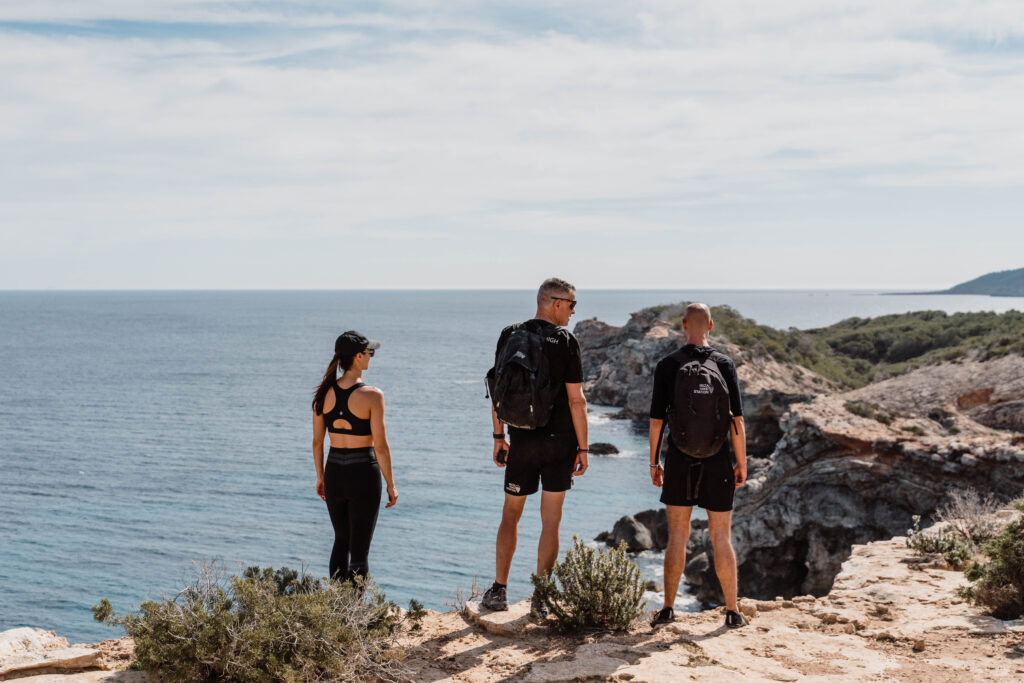 Three hikers pause on a rocky Ibiza coastal trail overlooking the sea.