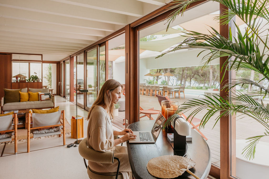 A guest works on a laptop at a table beside the villa’s glass doors and terrace.