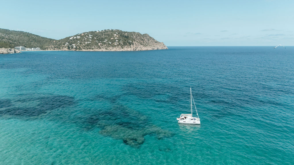 A sailboat floats in clear turquoise water off Ibiza’s rocky coastline.