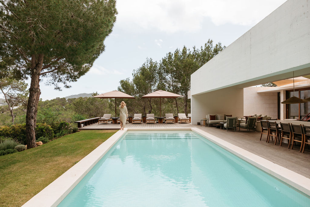 View along the villa pool toward a guest, shaded loungers and outdoor dining area.
