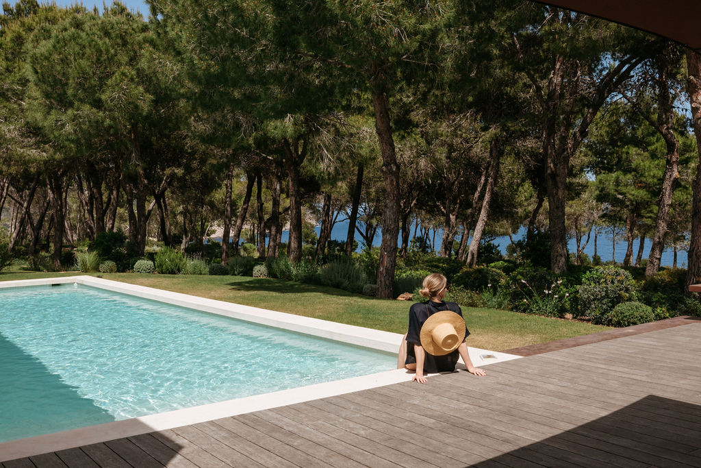A guest relaxes beside the villa pool, shaded by pine trees with the sea beyond.