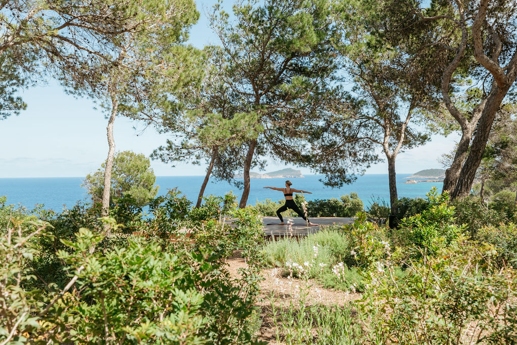 A guest practices yoga on a garden platform among pine trees overlooking the sea.