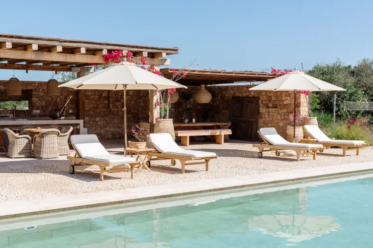 Rustic poolside terrace with beige umbrellas, wooden loungers, rattan chairs, hanging lanterns, stone wall, pink bougainvillea, and outdoor dining setup in sunny villa