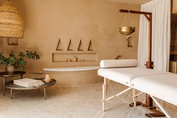 Minimalist Mediterranean spa interior with beige plastered walls, massage table, hanging rattan lamp, wooden shelves with pottery and incense, curved benches, and neutral earthy tones