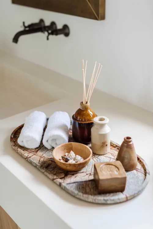 Rustic wooden tray with rolled white towels, reed diffuser, seashells, handmade soap, small pottery jars, and bronze faucet in spa bathroom setting