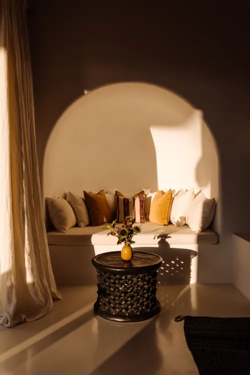 Intimate arched alcove seating with beige built-in bench, layered mustard and neutral pillows, dried flower arrangement in yellow vase, black woven table, sheer curtains, and warm golden lighting in rustic stone room