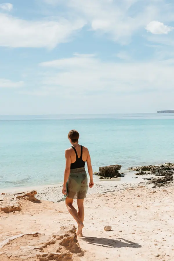 Marcia in a swimsuit and sarong walking onto the beach on Playa Migorn
