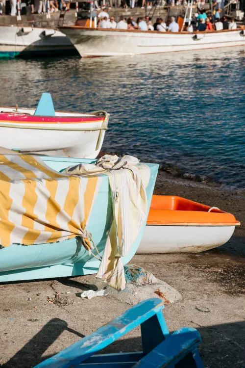 Boats resting by the shoreline; a blue and yellow striped cloth drapes over one. A crowd gathers on a dock in the background under the bright sun.