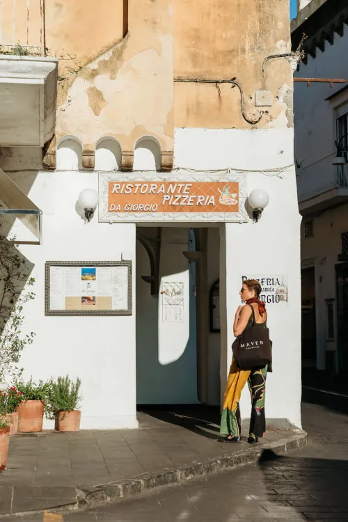 A woman stands outside a rustic Italian pizzeria, reading a menu on the wall. Sunlight casts warm shadows on the weathered building facade.
