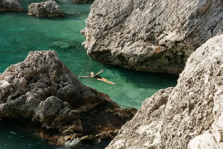 Person swimming in clear turquoise water surrounded by large rocky formations. The scene conveys tranquility and natural beauty, with sunlight casting soft shadows.