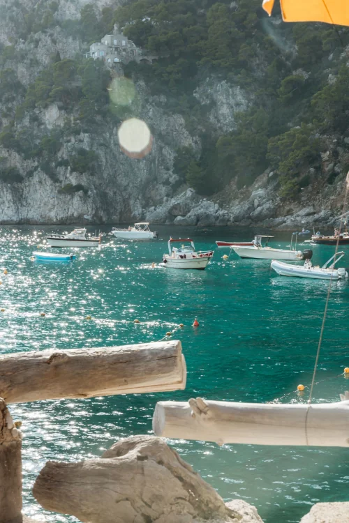 A sparkling turquoise sea with small boats sits against a backdrop of rocky, forested cliffs. Sunlight shimmers on the water, creating a peaceful vibe.