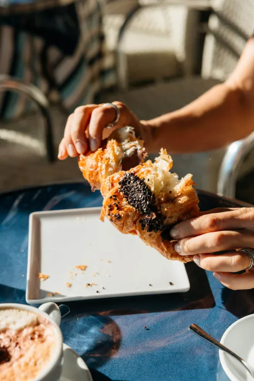 A person breaks a flaky chocolate croissant over a white plate on a sunlit cafe table. Nearby, there's a cup of frothy cappuccino, creating a cozy ambiance.