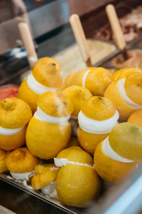 Lemons filled with creamy white gelato, nestled in a metal tray. The setting is colorful and inviting, suggesting a joyful, refreshing treat.