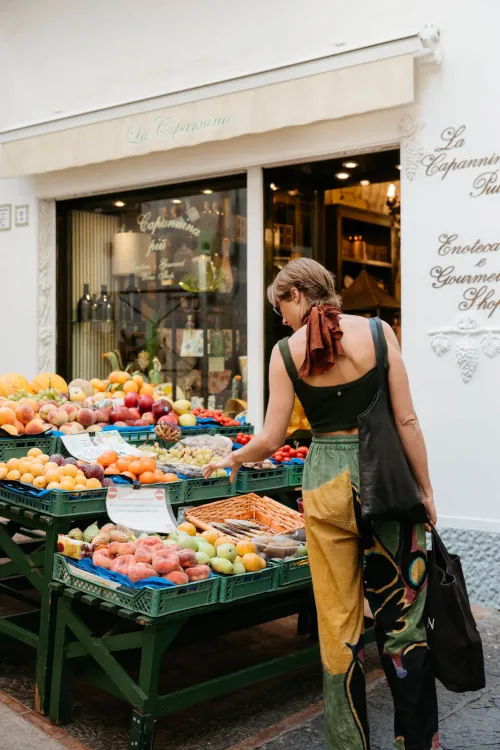 A person with a bag browses vibrant, fresh produce at an outdoor fruit stand beside a quaint shop. The scene is colorful and inviting.