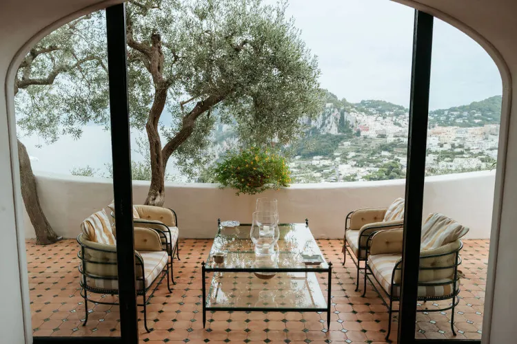Elegant patio overlooking a scenic hillside view, featuring four cushioned chairs around a glass coffee table atop a terracotta-tiled floor, creating a serene, inviting ambiance.