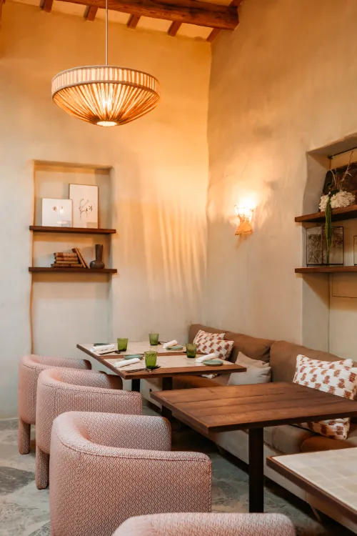 Cozy cafe interior with beige walls, a wooden table, cushioned chairs, and soft lighting. Shelves hold books and decor, evoking a warm, inviting atmosphere.
