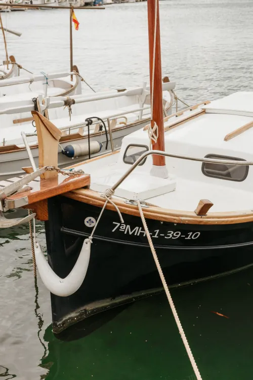 Close-up of a small white sailboat docked on calm water, featuring a wooden mast and mooring ropes. Nearby boats and a distant Spanish flag visible.
