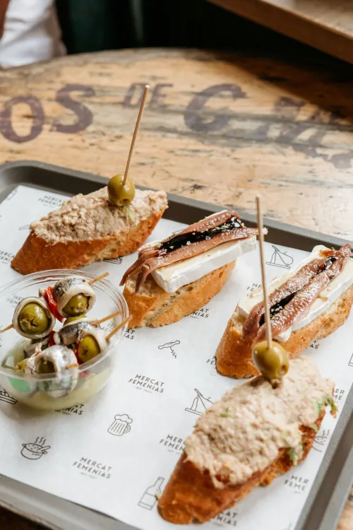 Toasted baguette slices with anchovies and cheese on a tray, garnished with skewered olives. A small cup of seasoned olives is beside, creating an inviting appetizer setting.