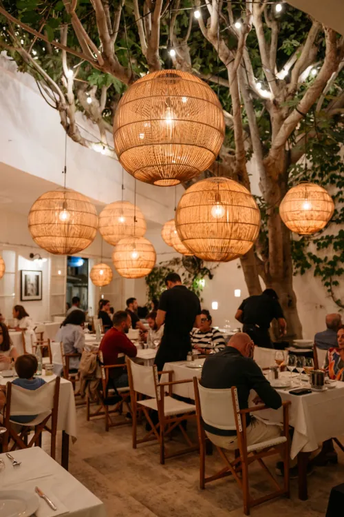 Cozy restaurant scene with large wicker pendant lights hanging from tree branches. Diners sit at tables with white tablecloths, creating a warm, inviting atmosphere.